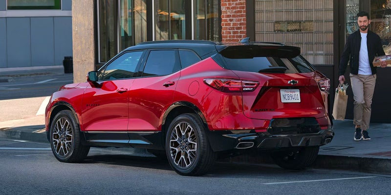 A red Blazer is parked in fron of a Building and a Person carrying Grocery moving towards Vehicle